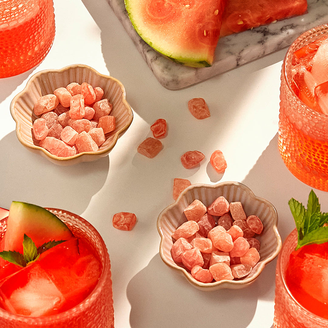 Mochi gummy candies presented in small beige scalloped bowls, with watermelon slices and pink drinks arranged around them on a bright white surface.